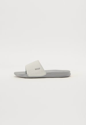 Jednopruhová šedá pantofle s děrovaným bílým páskem a logem „REEF“, mírně nakloněná doprava na jednoduchém bílém pozadí.