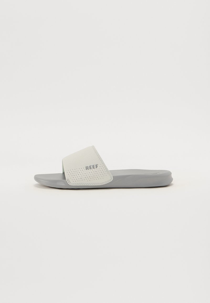 Jedna siva natikača s perforiranim bijelim remenom i logotipom "REEF", blago zakrenuta desno na jednobojnoj bijeloj pozadini.