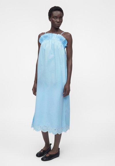 Γυναίκα που φοράει ένα ανοιχτό μπλε μίντι φόρεμα χωρίς μανίκια με βολάν στη λαιμόκοψη και κυματιστό τελείωμα, σε συνδυασμό με μαύρα παπούτσια Mary Jane.