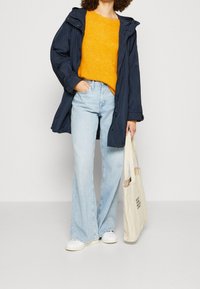Personne portant un imperméable navy, un pull orange vif, un jean large bleu clair, des baskets blanches, tenant un sac fourre-tout beige avec du texte.