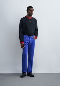 Homme debout contre un mur blanc portant une chemise noire à manches longues avec des poignets rouges, un jean bleu, des chaussures noires et un collier pendentif en or.
