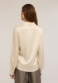 Crèmekleurige blouse met lange mouwen, een gladde textuur, een kraag, knoopmanchetten en een gebogen zoom, gedragen over gestreepte broek.