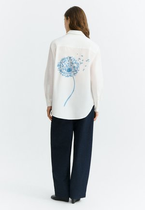 Vrouw met de rug naar de camera, draagt een wit shirt met een blauw paardenbloemontwerp op de rug en losse donkere broek tegen een egale achtergrond.
