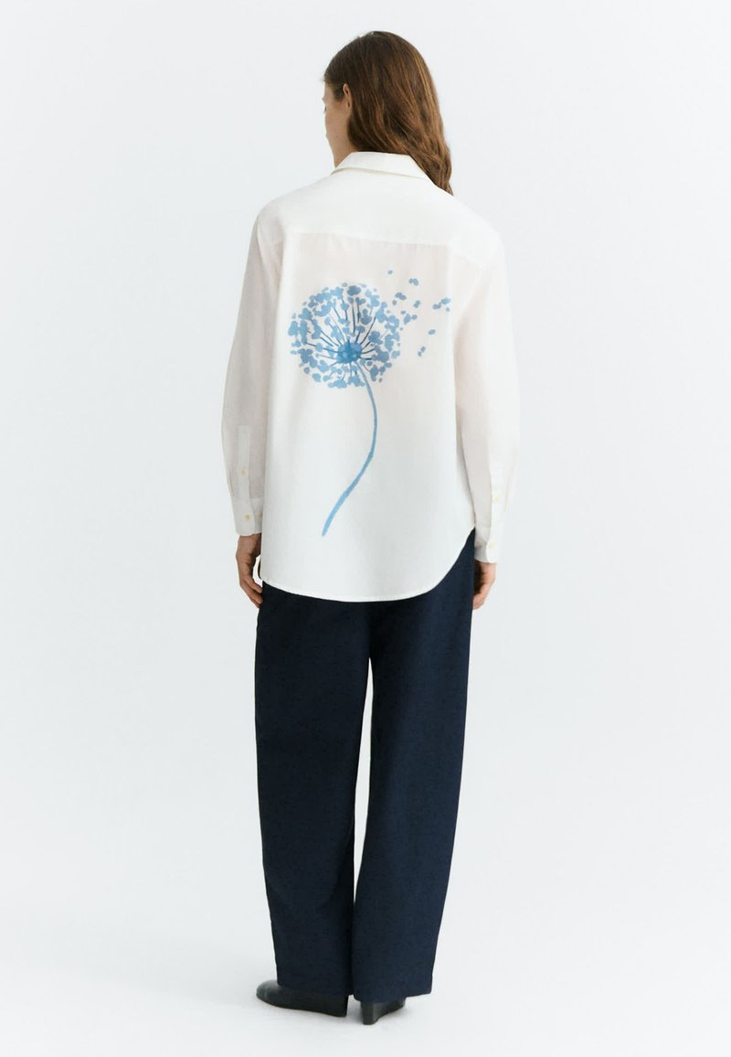 Femme vue de dos portant une chemise blanche avec un motif pissenlit bleu dans le dos et un pantalon ample sombre sur un fond uni.