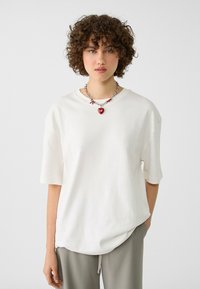 T-shirt bianca oversize realizzata in tessuto morbido con maniche corte. Collo tondo e vestibilità rilassata. Indossata con pantaloni grigi e una collana dettagliata.