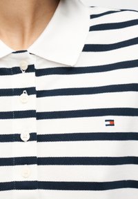 Κοντινό πλάνο ενός ατόμου που φοράει ριγέ πόλο μπλουζάκι σε λευκό και ναυτικό μπλε με λευκό γιακά και κουμπιά μπροστά.