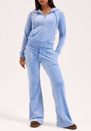 Donna che indossa una felpa con cappuccio in velluto azzurro chiaro e pantaloni larghi coordinati, con le mani nelle tasche, in piedi davanti a uno sfondo neutro.