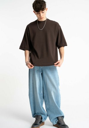 Młoda osoba w luźnych jasnoniebieskich dżinsach, oversize'owej ciemnobrązowej koszulce, łańcuszku i szarych trampkach, stojąca na białym tle.