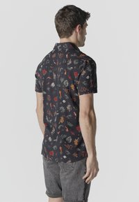 Hombre de espaldas lleva una camiseta negra de manga corta con un patrón colorido de peces koi, hojas y flores, combinada con shorts de mezclilla grises.