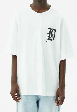 Camisa blanca de botones de manga corta hecha de algodón. Presenta un diseño de letra "B" negra contrastante en el lado izquierdo. Corte regular.