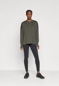 Suéter de manga larga verde oliva con hombros caídos, combinado con leggings negros y zapatillas negras. Textura suave, corte relajado.