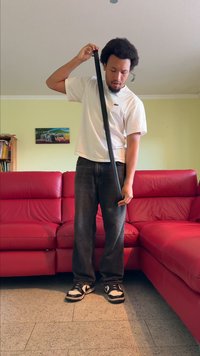 Henkilö pitää pitkää mustaa hihnaa, pukeutuneena valkoiseen paitaan ja tummiin farkkuihin, seisten teksturoitua lattiaa vasten punaisen nahkasohvan lähellä.