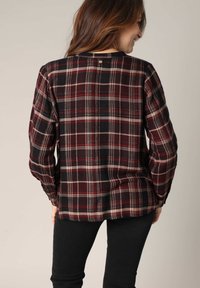 Chemise en flanelle à carreaux dans des teintes de rouge foncé, noir et crème. Elle présente des manches longues, un col arrondi et un détail de bouton au dos.