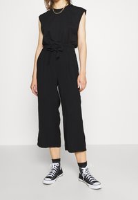 Combinaison noire sans manches avec taille ceinturée, pantalons larges et texture lisse, associée à des baskets montantes noires.