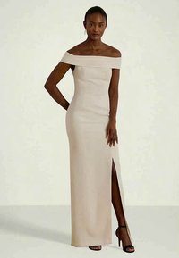 Vestido bege off-the-shoulder com uma silhueta ajustada, fenda lateral e textura suave, acompanhado por saltos pretos.