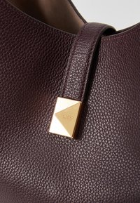 Tamsiai burgundi odinė rankinė su tekstūruotu paviršiumi. Pabrėžia auksinę trikampio formos detalę su įspaudų logotipu. Lygaus paviršiaus pamušalas.