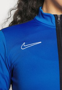 Blauwe sportjack met hoge kraag, ritssluiting en zwarte accenten. Witte Nike-logo geborduurd op de bovenste linkerborst.