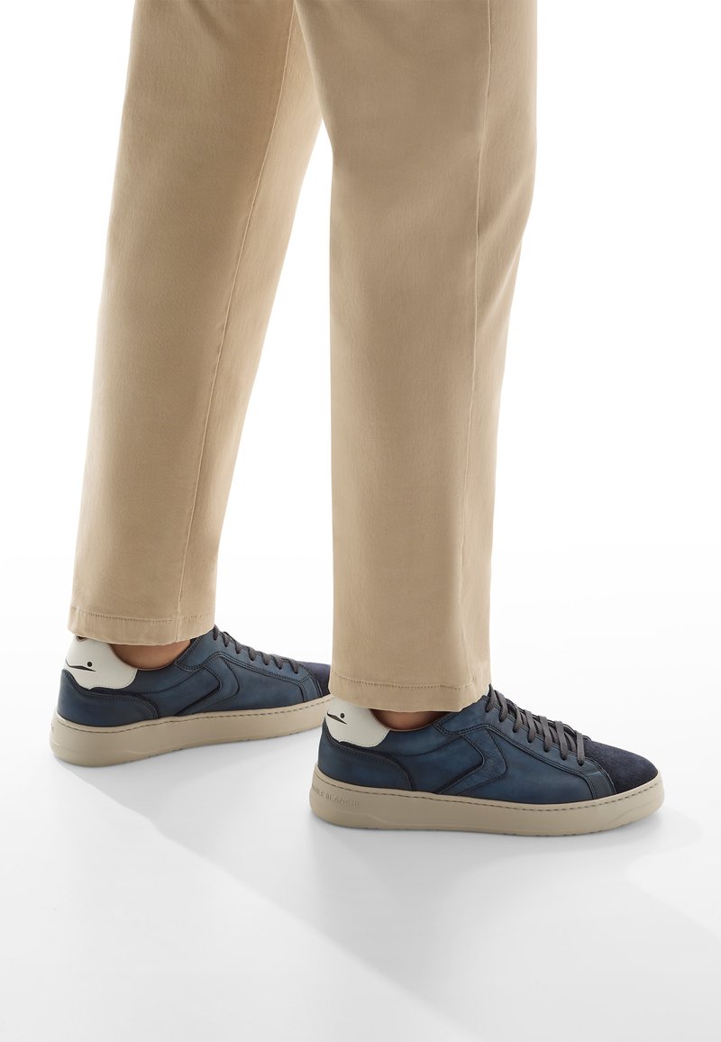 Persona che indossa pantaloni beige e sneakers basse blu scuro con dettagli bianchi sul tallone, in piedi su una superficie bianca.