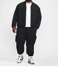 Svart lätt bomberjacka med dragkedjefickor över en vit t-shirt, kombinerad med svarta cargo-byxor och vita sneakers med svarta accenter.