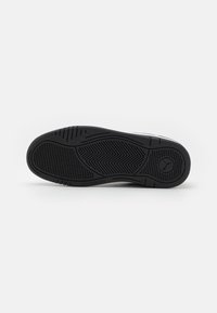 Semelle de chaussure noire avec motifs texturés pour une meilleure adhérence et un logo circulaire près du talon sur fond blanc uni.