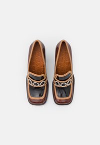 Mocassins en cuir verni marron et noir avec des bordures beige et un détail en chaîne de strass, vus de dessus sur un fond blanc.