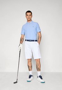 Ανοιχτό μπλε πόλο μπλουζάκι με εφαρμοστή γραμμή, άσπρες σορτς και μπλε λεπτομέρειες. Άσπρα παπούτσια με σκούρα μπλε σόλα και ρόπαλο του γκολφ στο χέρι.