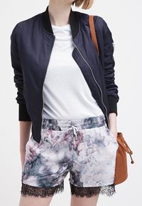 Námornícka bomber bunda se zipem, bílý tričko a květinové šortky s krajkovým lemem. Hnědá taška s popruhem dotváří celý outfit.