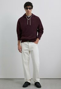 Sudadera burdeos con cordones, camiseta gris debajo, pantalones anchos blancos y zapatos negros, acompañados de gafas de sol negras.