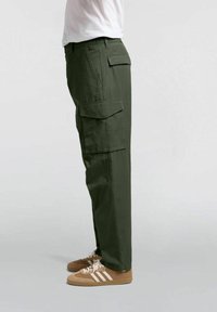 Pantaloni cargo verde oliva con molteplici tasche laterali, design a gamba dritta e tessuto leggero. Indossati con scarpe da ginnastica beige.