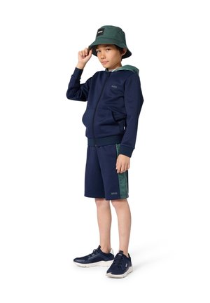 Survêtement bleu marine avec un sweat à capuche zippé et un short avec des accents verts. Associé à un bob vert et des chaussures de sport bleu marine.