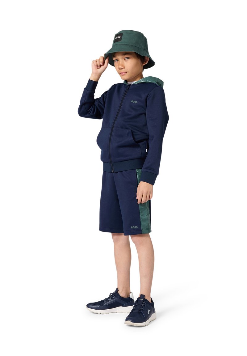 Chándal azul marino con una sudadera con capucha y cremallera, y pantalones cortos con detalles en verde. Combinado con un sombrero tipo pescador verde y zapatillas deportivas azul marino.