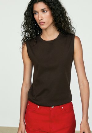 Femme aux longs cheveux bouclés portant un haut marron foncé sans manches et un pantalon rouge vif, debout devant un fond clair uni.