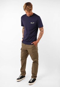 T-shirt bleu marine à col rond avec un petit logo multicolore. Pantalons cargo olive avec plusieurs poches, portés avec des baskets noires.
