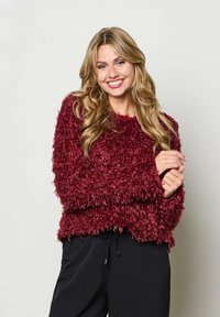 Bordeaux fuzzy trui met een cropped ontwerp, ronde halslijn en lange mouwen. De textuur lijkt zacht en harig. Draagt over zwarte broek.