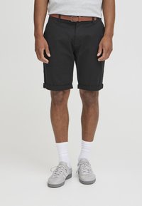 Svarte bomullsshorts med rullekanter, parret med et brunt belte. Modellen bruker grå sneakers og hvite sokker. Enkelt, uformelt design.