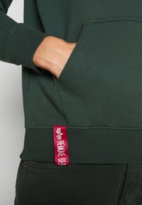 Zöld pamut kapucnis felső bordás mandzsettákkal, kenguruzsebb és egy piros címke a szegélyen, fehér szöveggel és grafikával.