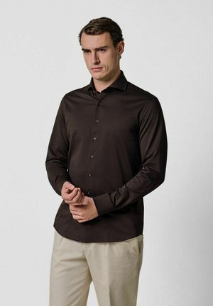 Brązowa koszula z długim rękawem wykonana z gładkiej tkaniny o dopasowanym kroju, z kołnierzykiem w szpic i zapinana na guziki z przodu, zestawiona z jasnymi spodniami.