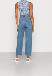 Jeans de tiro alto azul claro con piernas anchas y un diseño de bolsillo trasero de estilo vintage. Combinados con un top floral ajustado y sandalias blancas.