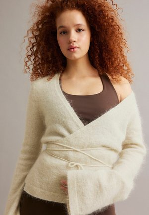 Młoda kobieta o kręconych rudych włosach, ubrana w kremowy, puszysty sweter typu wrap na brązowej koszulce na ramiączkach, stojąca na neutralnym tle.