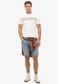 Camiseta blanca de algodón con rayas horizontales de colores y texto "SuperDry", combinada con pantalones cortos de mezclilla azul y una camisa a cuadros roja atada en la cintura.