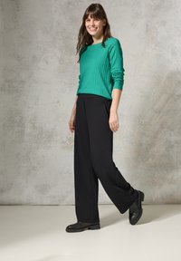 Grüner gerippter Langarmpullover kombiniert mit hochgeschnittenen schwarzen weiten Hosen und schwarzen Knöchelstiefeln, vor einem neutralen Hintergrund.