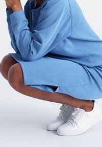 Personne accroupie portant une robe bleue à manches longues avec des fentes latérales et des baskets blanches à lacets sur fond blanc.