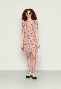 Vestido rosa com padrão floral, mangas curtas, decote profundo e saia rodada. Combinado com sandálias brancas e uma bolsa bege.