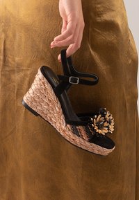 Sandalia de cuña negra de ante con base de yute trenzado, que presenta un acento floral y una correa en el tobillo. La textura es suave con un acabado similar al ratán.