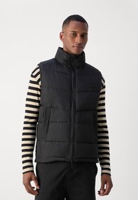 Black puffer vest with high collar, zip closure, and side pockets. Worn over a striped long-sleeve shirt, featuring a smooth texture.