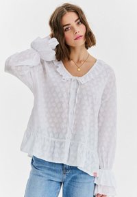 Biała, teksturowana bluzka z falbankowym kołnierzykiem i rękawami, zapinana na guziki z przodu, z falbanowym dołem. Zestawiona z jasnoniebieskimi dżinsami.