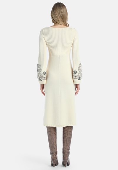 Ženska v kremnem midi obleki s rebrastim pletenjem in vezenimi rokavi, v kombinaciji z visokimi teksturiranimi rjavimi škornji, na enobarvni beli podlagi.
