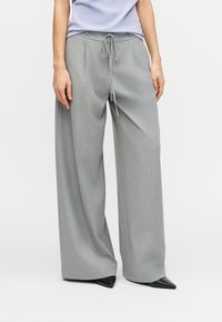 Kvinde iført lysegrå bukser med vidde i benene og snoretræk i taljen samt sorte spidse sko mod en ensfarvet baggrund.