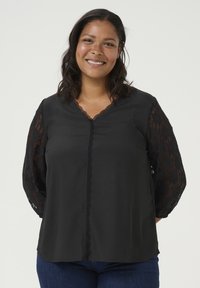 Black blouse featuring a V-neck with lace accents and long lace sleeves, paired with dark denim jeans. Smooth fabric with a structured fit.