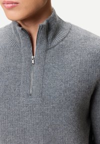 Nahaufnahme einer Person, die einen grauen, gerippten Feinstrickpullover mit einem halben Reißverschluss aus Metall und einem hohen Kragen trägt.